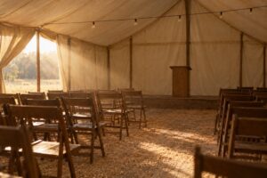 The empty interior of an early twentieth-century revival meeting tent, rows of wooden folding chairs on a sawdust floor in late afternoon light